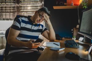 An unhappy man seated at a computer looks at his phone and wonders if it is possible to recover lost crypto.