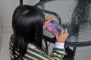 Young girl with long dark hair, seen from behind, holding a smartphone sideways and playing a colorful 3D game at a glass patio table