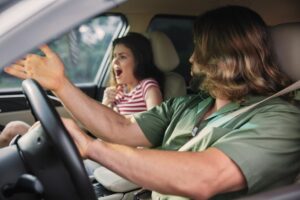 Frightened woman screaming in the front passenger seat of a car while the male driver turns toward her with his hand raised