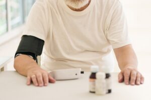 Older man with a gray beard wearing a blood pressure cuff on his arm, seated at a table with a home monitor and medication bottles