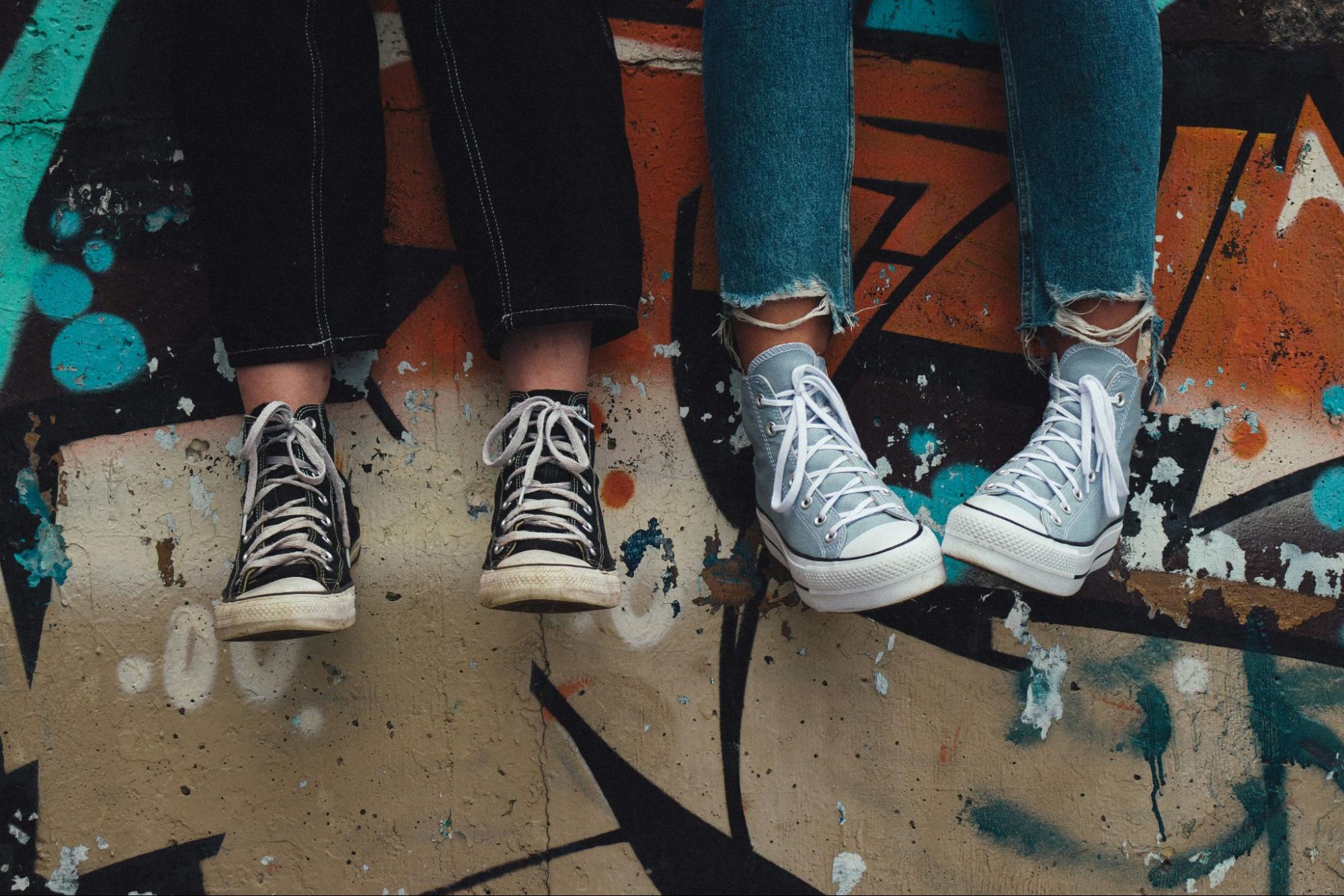 Two young people sitting on a graffiti covered wall, one wearing black high top sneakers and the other light blue high tops with ripped jeans